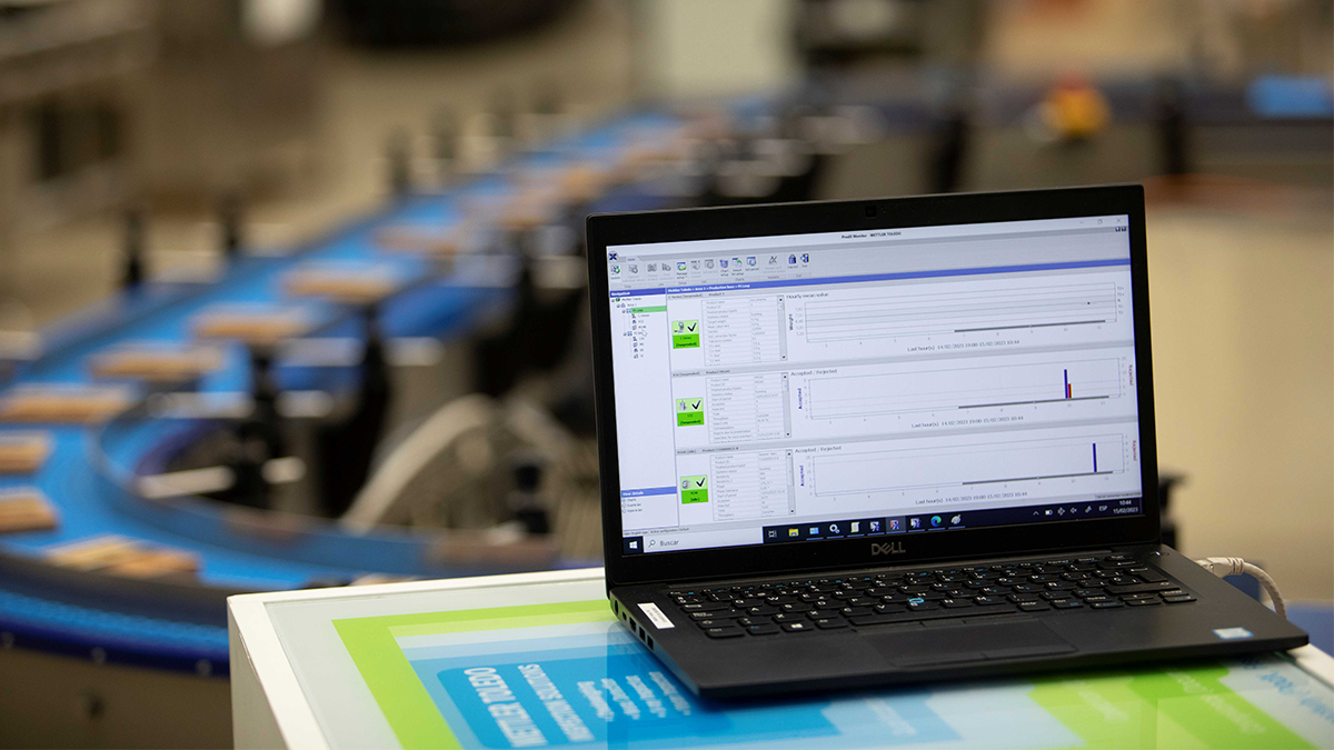 A close-up of a laptop displaying software used to control or monitor a production line, with parts of a conveyor belt and additional machinery visible in the background. The image conveys a sense of a modern, digitally monitored production environment.