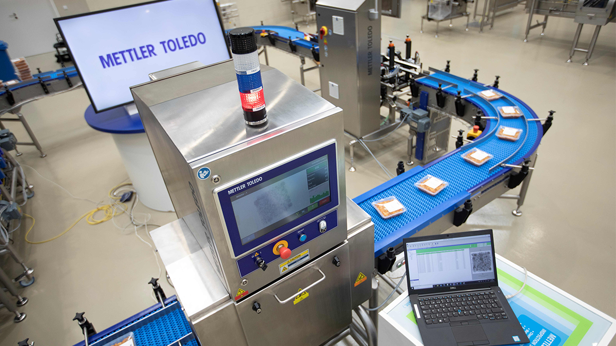 Overview of an automated production line at METTLER TOLEDO: A conveyor belt transports packaged products, and in the foreground is a machine with a digital display and an alarm signal. The machine's screen shows software for monitoring the production process.