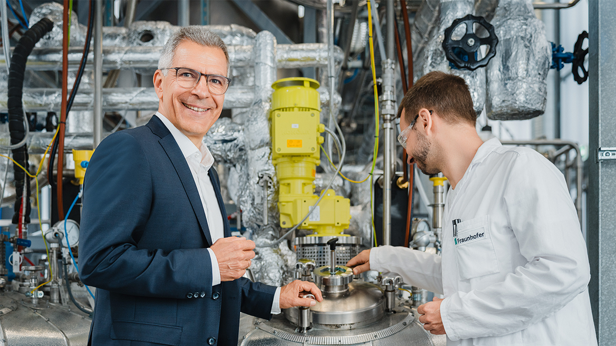 Two men are in a technical or industrial setting. One is wearing a dark business suit with a white shirt, the other a white lab coat. They appear to be having a discussion or conducting an inspection. The man in the lab coat is actively working on a round metallic device or reactor, while the suited man listens or asks questions. In the background, there is a complex arrangement of pipes, valves, and technical components indicating a highly advanced facility.