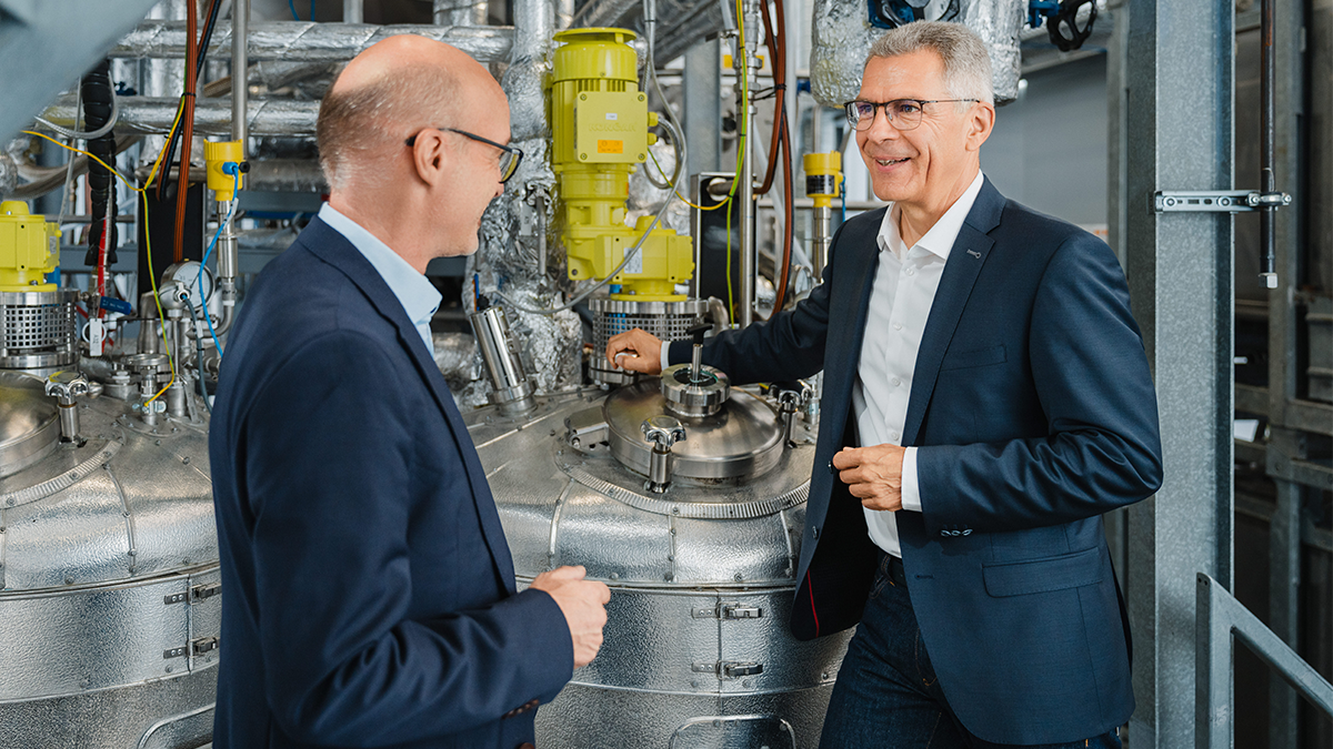 Two men in dark suits are in a highly technical environment with numerous pipes, valves, and additional technical elements. They are engaged in a discussion, likely during a tour or technical inspection. The man with a white shirt and dark suit seems to be the host or person in charge, while the other is also inspecting the facility.
