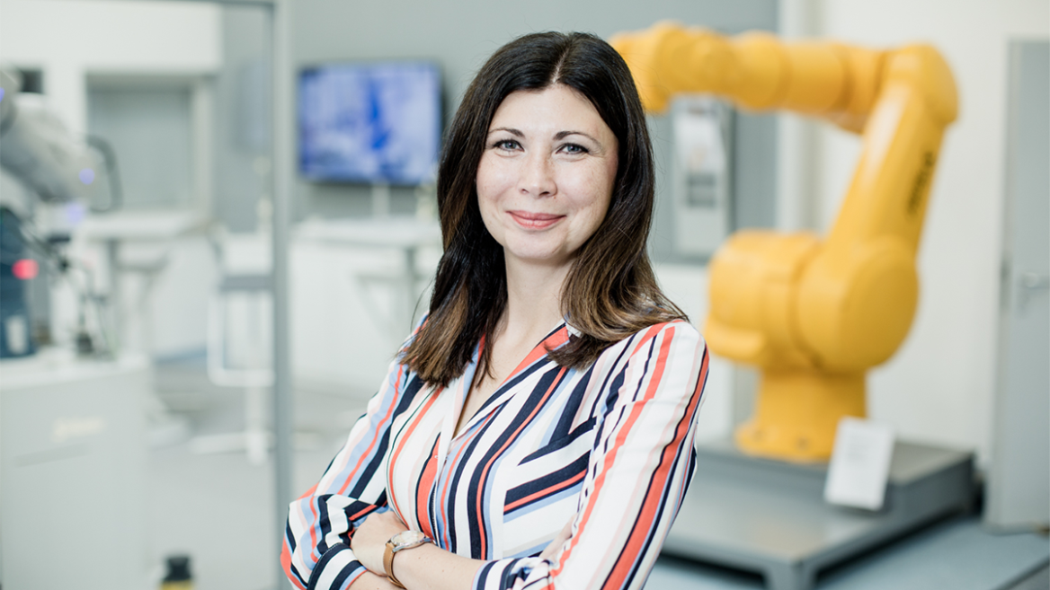 Eine Frau arbeitet in einem modernen Labor, trägt eine gestreifte Bluse und hat die Arme verschränkt. Im Hintergrund sind technische Geräte und ein automatischer Roboterarm sichtbar, was auf eine High-Tech-Umgebung in Forschung oder Entwicklung hindeutet.