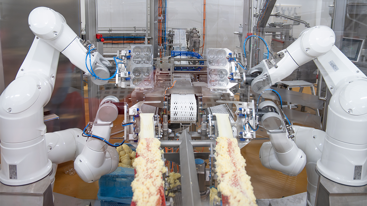 Eine komplexe Robotik-Anlage mit mehreren Armen und Greifern arbeitet in einer Industriehalle. Die Maschine scheint für Montage- oder Verpackungsaufgaben eingesetzt zu werden.