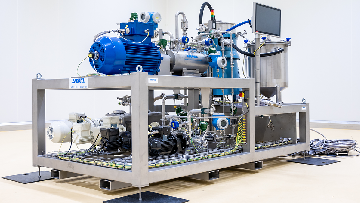 Eine große technische Maschine, vermutlich für die Wasser- oder Luftaufbereitung oder eine andere industrielle Anwendung, besteht aus zahlreichen Rohren, Pumpen, Motoren, Messgeräten und weiteren Komponenten auf einem stabilen Metallrahmen. An der Maschine ist ein Monitor montiert. Die helle, gut beleuchtete Umgebung erinnert an eine Industriehalle oder einen Reinraum.