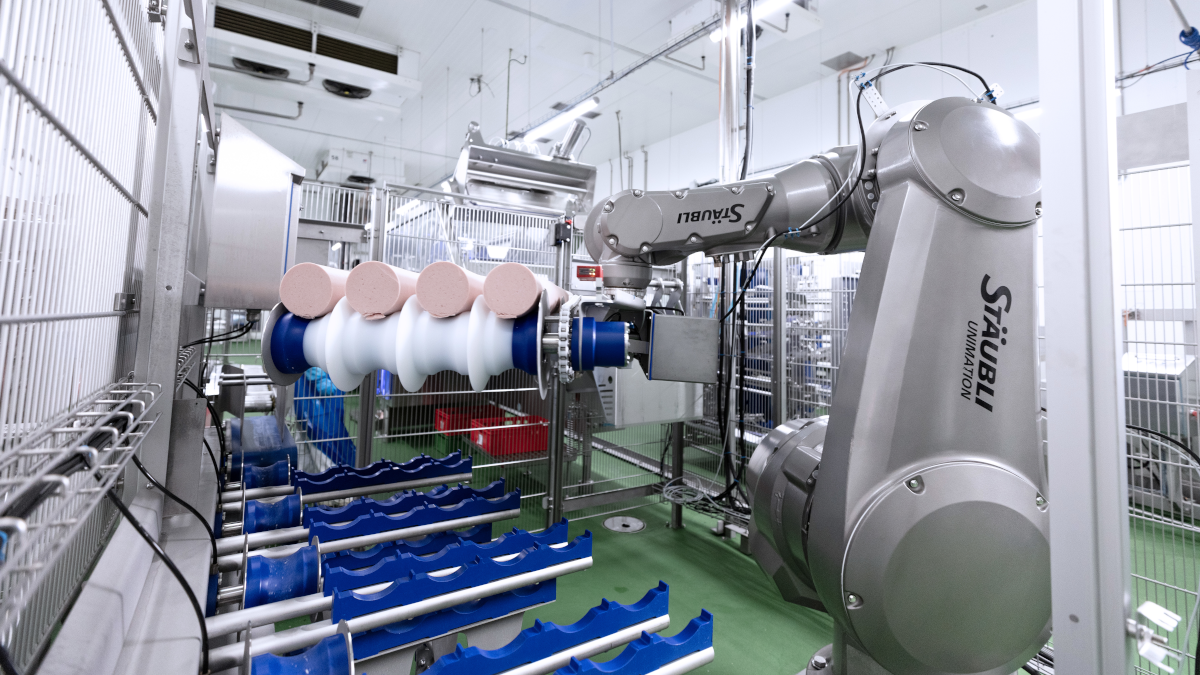 A highly automated production facility featuring a robot-like system holding an orange, white, and blue product on a rotating roller or conveyor belt. The environment is marked by metal fencing and technical components, producing a sterile, controlled atmosphere.