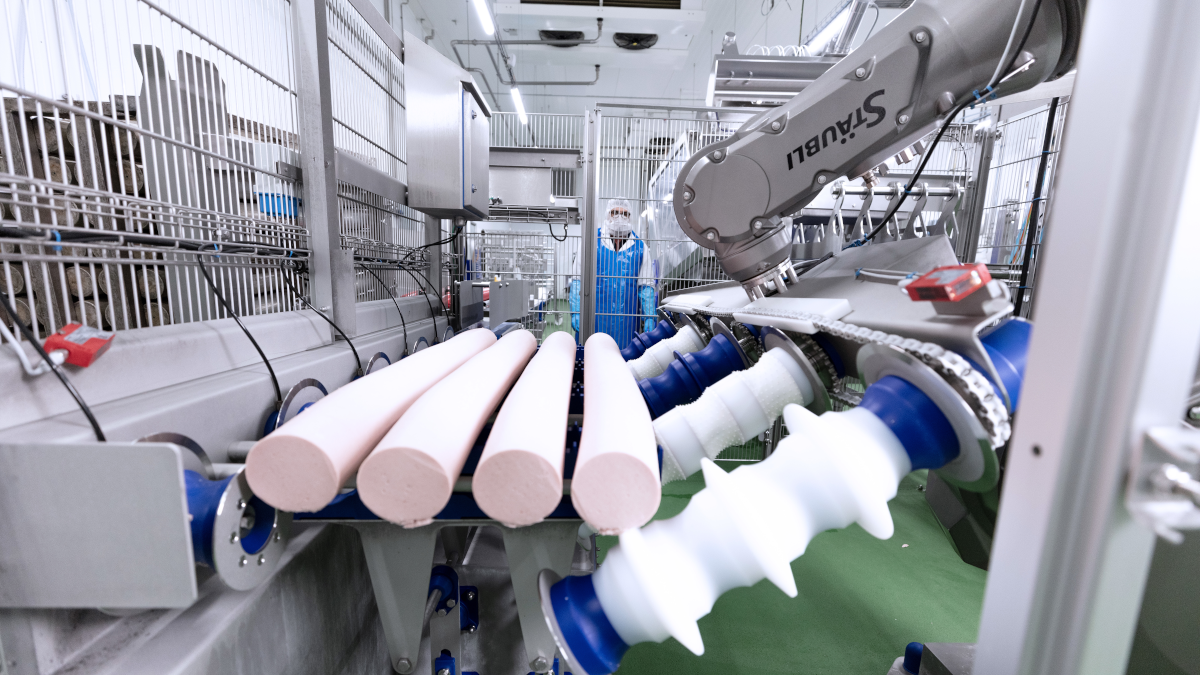 Several red, round objects rest on a conveyor belt, with a human operator wearing a protective mask visible in the background. The scene displays another part of the automated facility in a clean, structured setting.