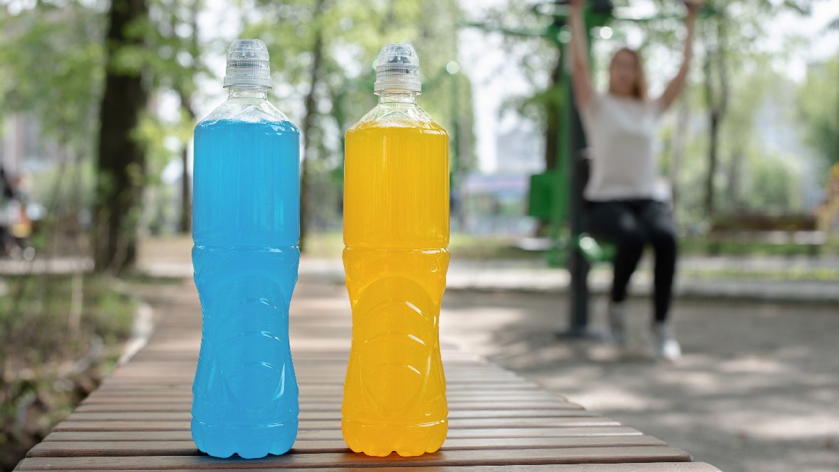 Two bottles of blue and yellow drink stand on a wooden table and a woman trains in the background.