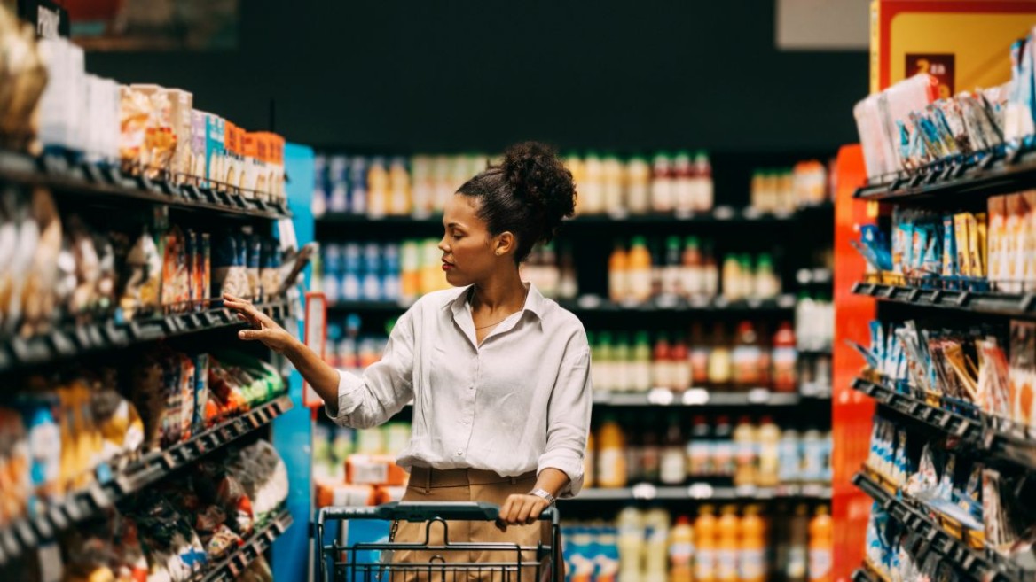 A woman is standing in a supermarket with a shopping trolley, looking at different products.