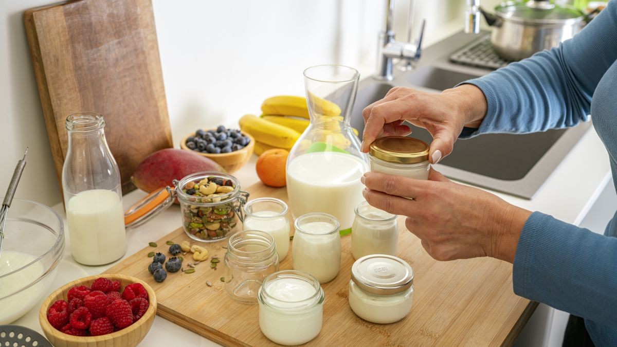 Person opening a jar of yogurt on a kitchen counter with milk bottles, fruit, and nuts.