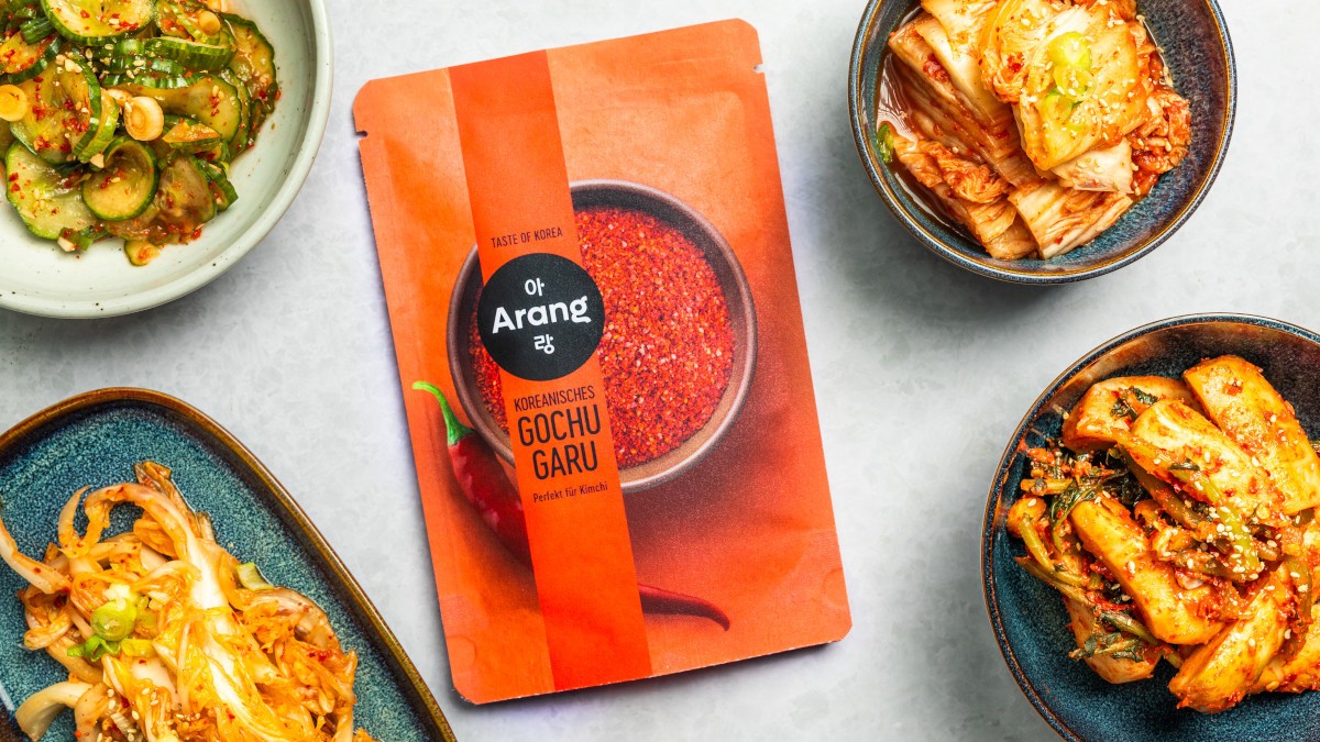 Eine Tüte mit Gochugaru von Arang umringt von Schüsseln mit gewürzten Lebensmitteln wie Kimchi oder Gurken – repräsentativ für die koreanische Küche. 