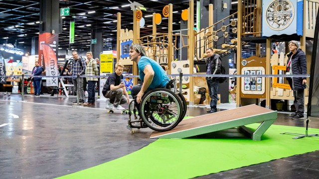 A person in a wheelchair rides a ramp from Street Unit in the SkateArea at FSB 2025. Play equipment and people can be seen in the background.