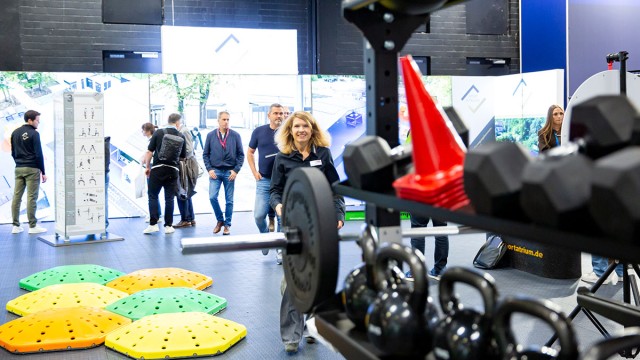 Messestand von SportAtrium mit Trainingsgeräten wie Kettlebells, Hantelscheiben und bunten Balance-Pads, mehrere Personen im Hintergrund.