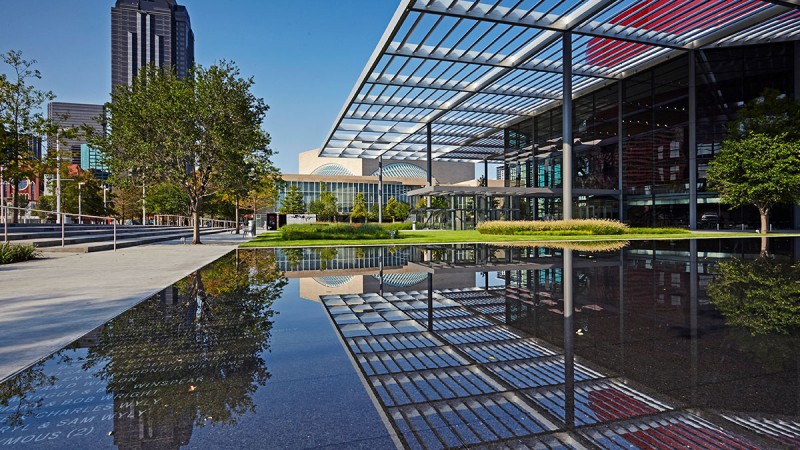 Moderne Architektur mit Glasfassade und reflektierender Wasserfläche im Vordergrund. Bäume und Sträucher umgeben das Gebäude.