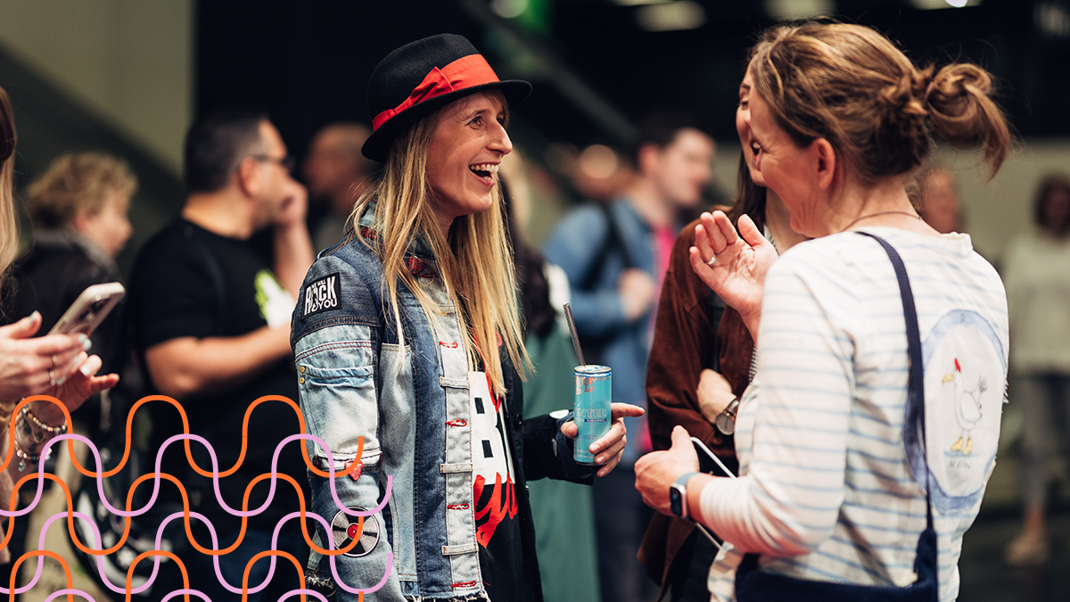 Zwei Frauen im Gespräch, eine trägt Jeansjacke und Hut, die andere gestreiftes Oberteil und Rucksack, im Hintergrund mehrere Personen.