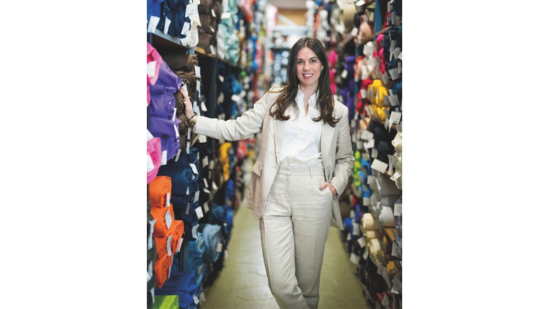 Frau in hellem Anzug steht in einem Gang mit Regalen voller bunter Stoffrollen, eine Hand an einem Regal.