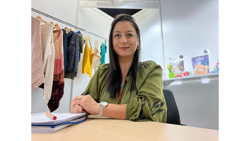 Frau mit langen schwarzen Haaren und grüner Bluse sitzt an einem Tisch mit Notizbuch, im Hintergrund hängen gestrickte Kleidungsstücke an einer Stange