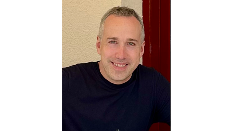 Man with short gray hair wearing a black t-shirt sitting in front of a light wall.