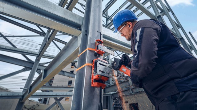 Ein Arbeiter mit Schutzhelm und Arbeitskleidung verwendet einen FEIN VersaMAG an einem Metallträger, während er das Werkstück mit einem Winkelschleifer bearbeitet.
