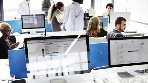 mehrere Personen in einem großen Büro mit Tischen, auf denen Bildschirme stehen