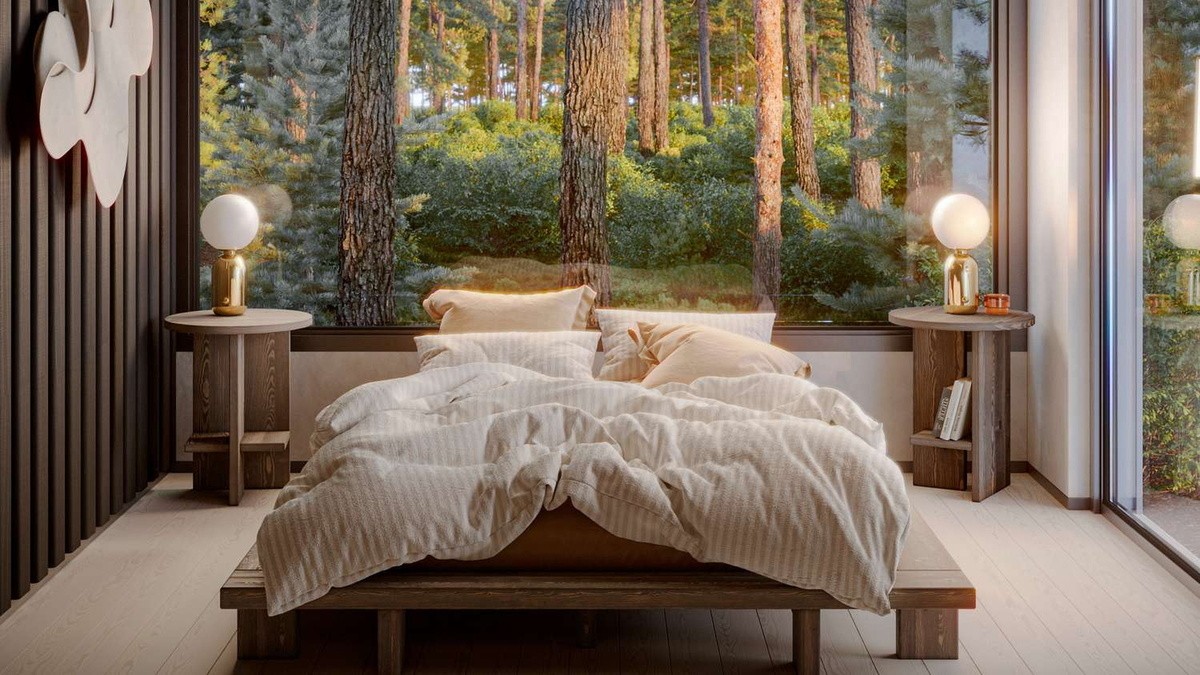 Bedroom with a wooden bed positioned in front of a large window overlooking a forest. The bed features cream-colored bedding and pillows; nightstands hold lamps with round white shades and small decorative items. The room exudes a warm, natural atmosphere with plenty of wood and natural light.
