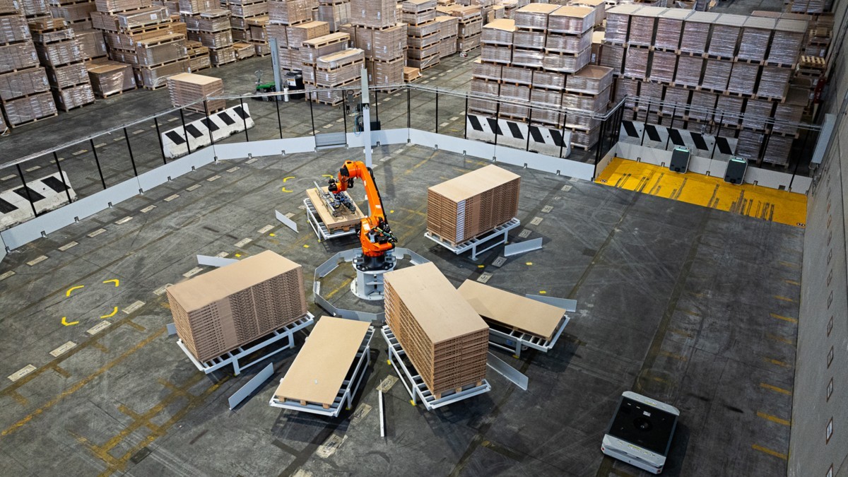 A large industrial warehouse with a robotic arm in the center handling wooden panels on multiple pallet stacks. Surrounded by tall shelves filled with wooden products. The floor is marked for logistical operations.