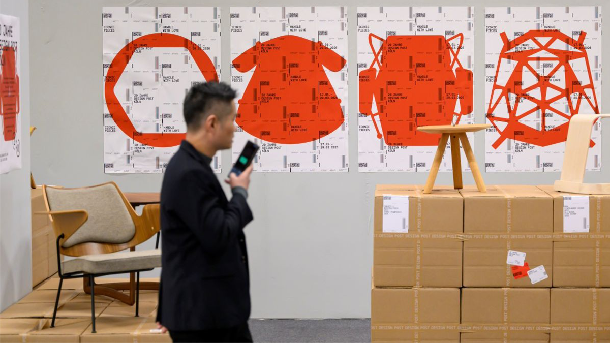 Impression von der imm cologne: Messebesucher mit Smartphone vor einer Wand mit roten Grafiken, daneben Stuhl, Hocker und gestapelte Kartons.