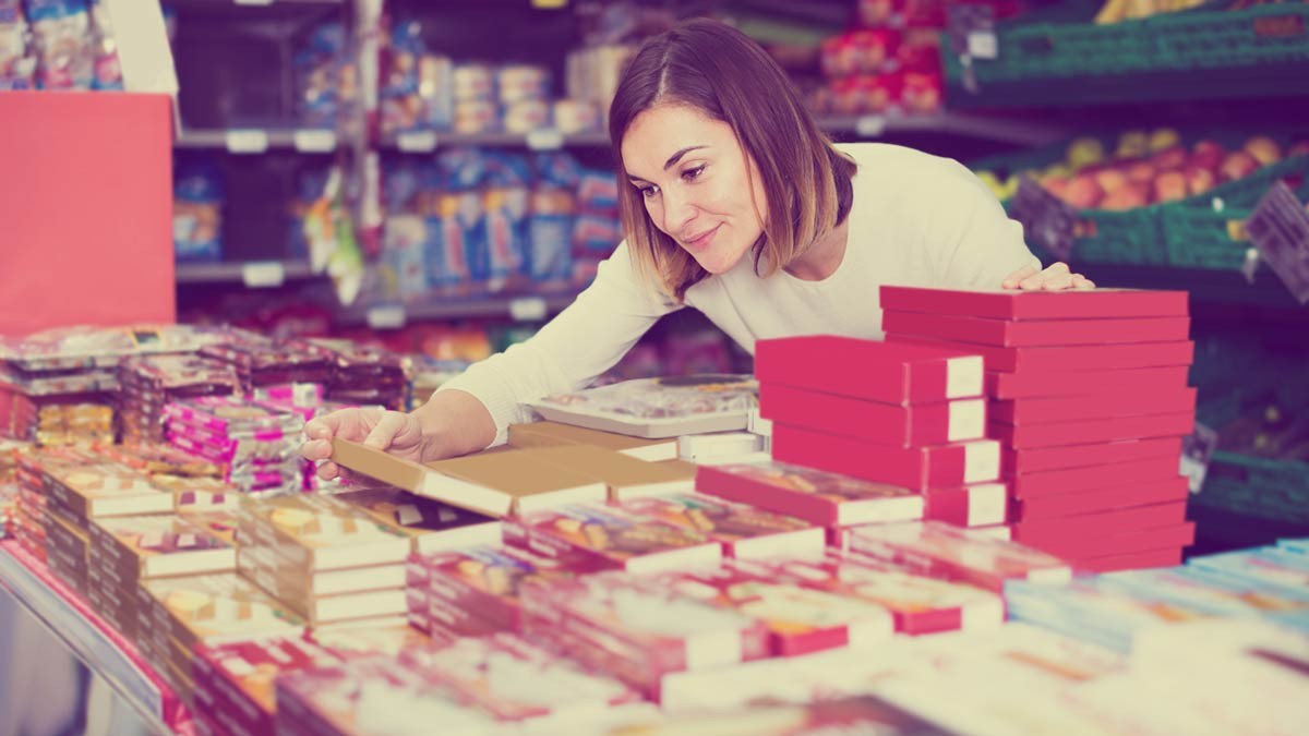 A woman leans over the confectionery selection in a shop and smiles as she looks at a package.