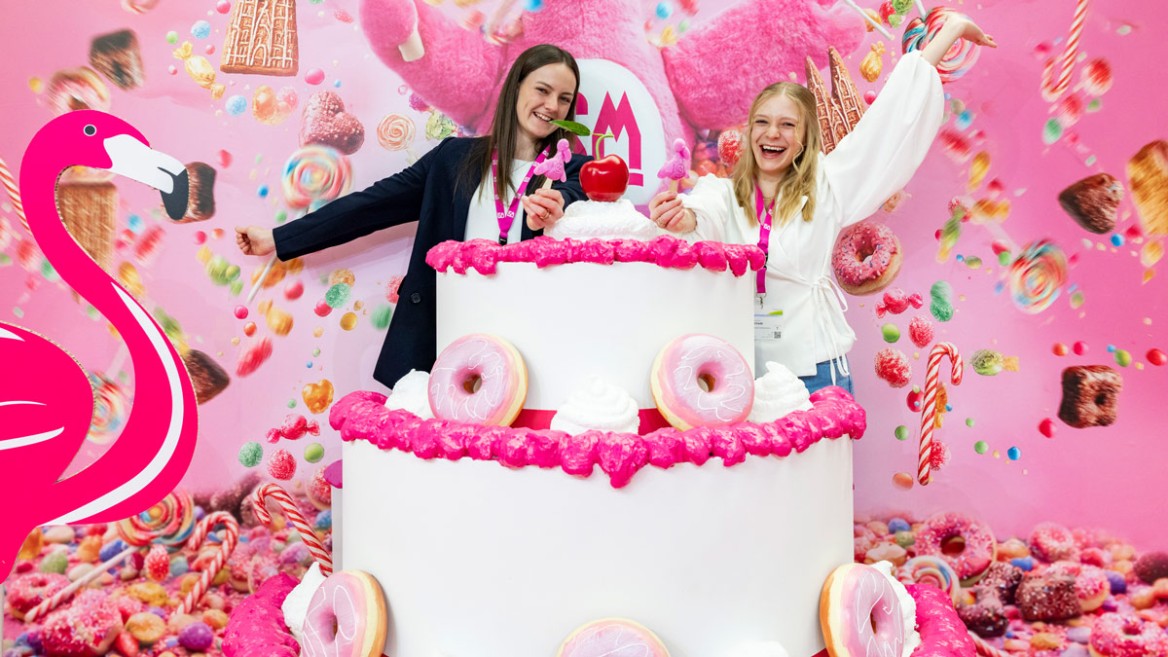 Two people posing with the ISM mascot GISMO at the photo point behind a huge cake at ISM 2026, the hashtag #ISMfamily can be seen in the background.