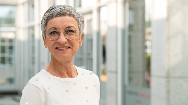 Person with short gray hair wearing a white top with small decorative elements, standing in a modern office building corridor