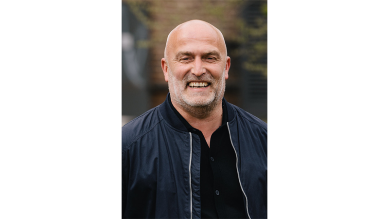 Bald man wearing a black jacket over a black shirt, standing outdoors with blurred background