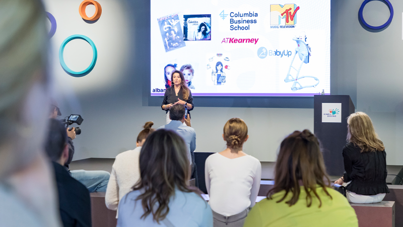 A woman is giving a presentation in a modern conference room in front of a large digital screen displaying various logos such as MTV, Columbia Business School, BabyUp, and AT Kearney. The audience sits attentively on chairs, and one person is holding a camera to record the talk.