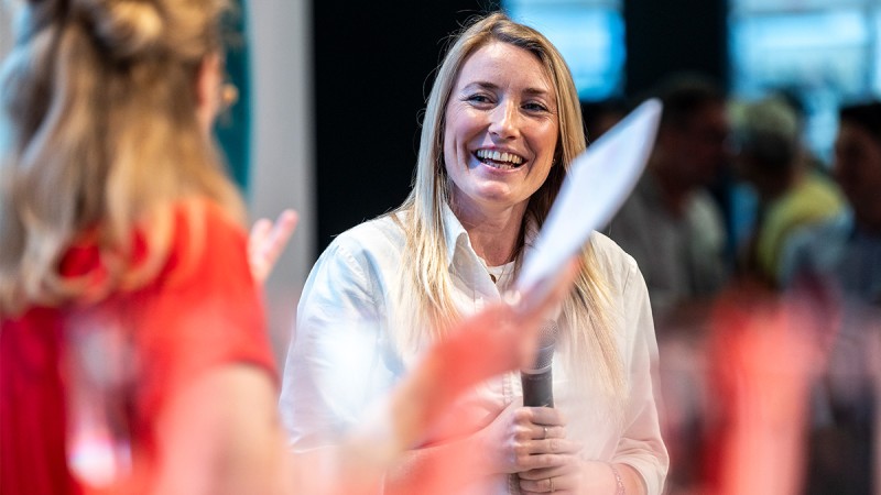 A woman in a white shirt is speaking into a microphone in front of a group of listeners. The image is blurry, focusing on the woman, likely during a discussion or Q&A session at an event.