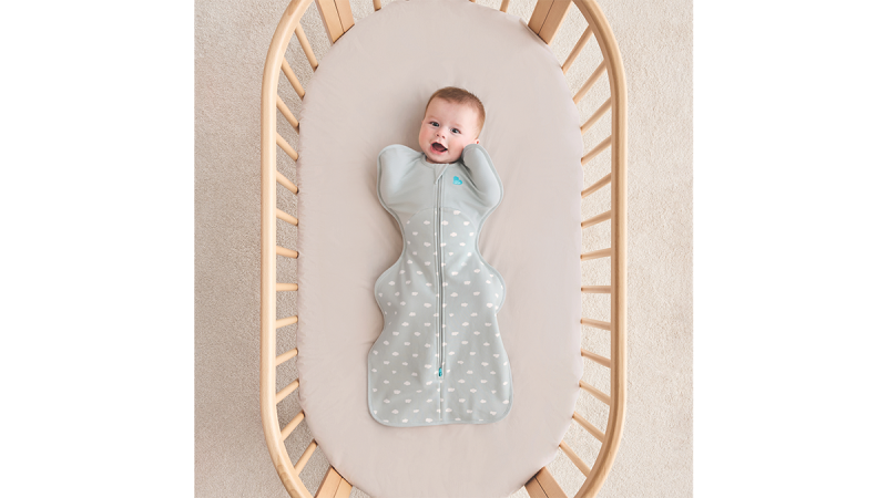 Baby lying on back in a wooden crib wearing a gray Swaddle Up Pucksack with white dots and a front Zipper
