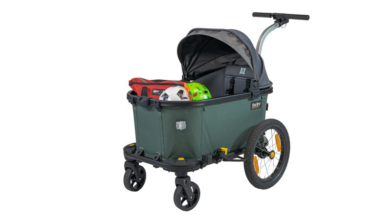 Burley cargo wagon with green fabric container, black canopy, two large rear wheels, and two small front wheels, carrying a red bag and a green helmet