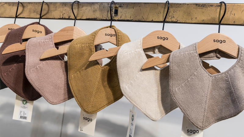 A row of baby hats or bibs in neutral natural tones such as brown, beige, olive green, and gray are neatly hung on wooden hooks on a coat rack.