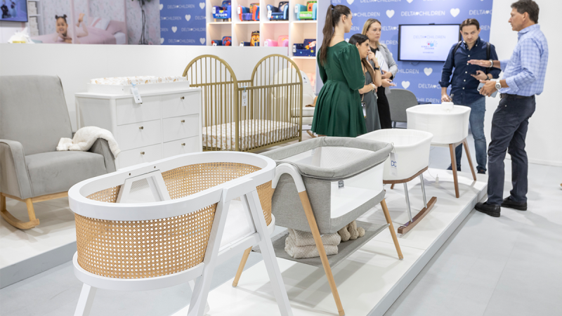 An exhibition display of children's furniture at Kind und Jugend. Various baby seats, including a high chair, as well as different children's beds and cradles are arranged on a platform. In the background, several people are talking or exploring the displayed products. The scene conveys the atmosphere of a presentation of innovative baby seats and children's room furniture at Kind und Jugend.