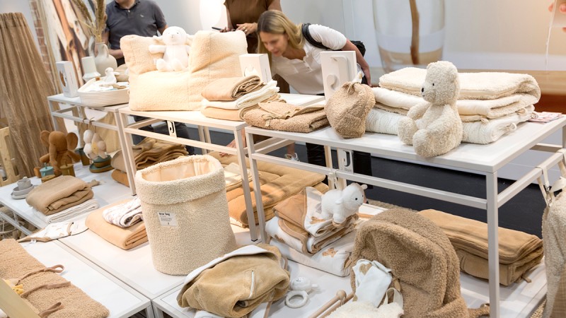 A small selection of baby and children’s textiles, such as towels, bibs, and washcloths in warm, neutral tones, as well as some small fabric toys, are laid out on a white table in the store.