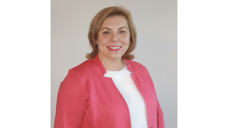 Woman with shoulder-length blonde hair wearing a coral blazer over a white top, standing against a plain background