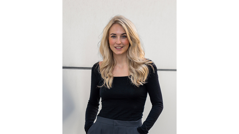 Person with long wavy blonde hair wearing a fitted black long-sleeve top and dark gray pants, standing with hands in pockets