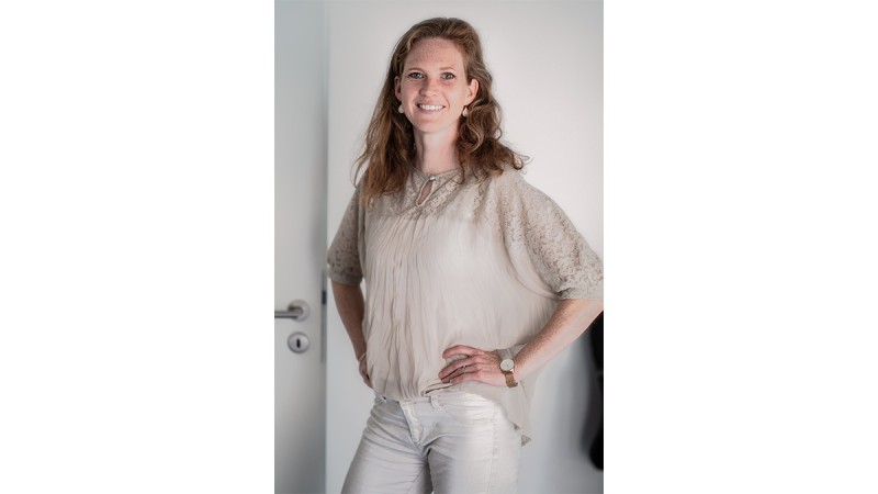Woman with long wavy hair wearing a beige lace blouse and white pants, standing with hands on hips.