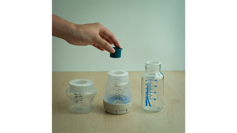 Hand placing a green cap on a white breast pump with a clear baby bottle and a milk storage container on a wooden surface