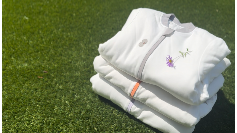 Stack of three white folded sweatshirts on grass, top one embroidered with a purple flower and green stem
