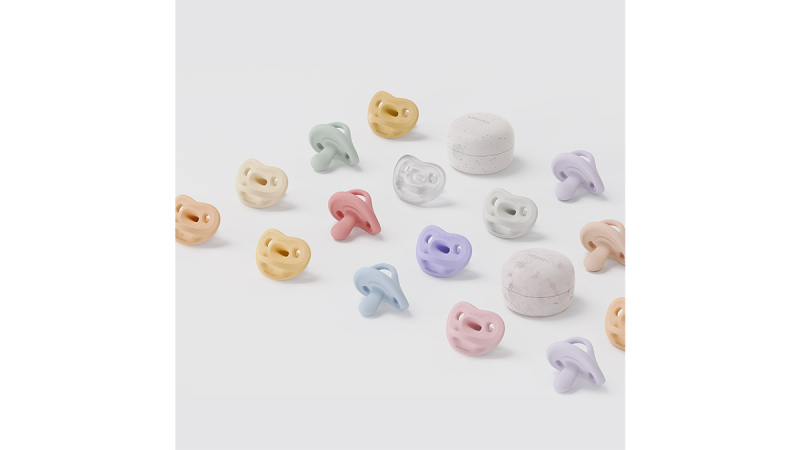 Multiple pastel-colored silicone pacifiers and two round white storage cases on a white surface