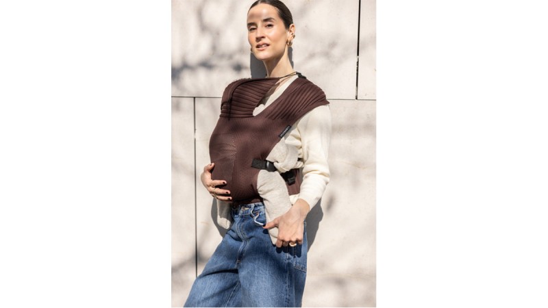 Person wearing a brown baby carrier with a baby inside, dressed in a cream sweater and blue jeans, standing against a light wall