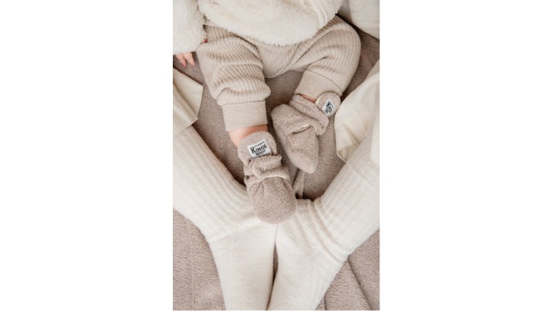 Baby wearing beige knit mittens and pants lying on adult's lap with white knit socks