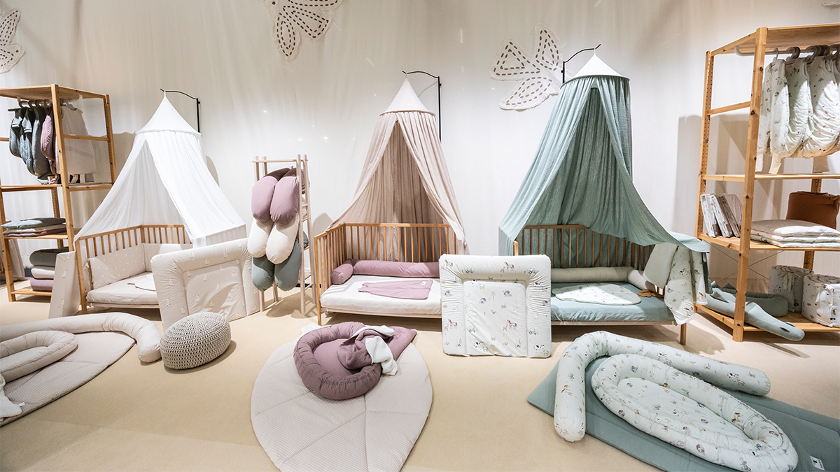 A lovingly decorated baby or child’s room featuring a wooden crib with blue and white striped bedding on the left side. In the middle, there is a light wood dresser with three drawers, topped with a small lamp giving off warm light, a picture frame, a toy car, a cup, and a box containing decorative items. A note is hanging from the dresser. On the right, there is a changing table with a beige changing pad and a cloth, next to a large plant with long, narrow leaves. Below the changing table is a round, woven basket. The overall atmosphere is bright, modern, and child-friendly.