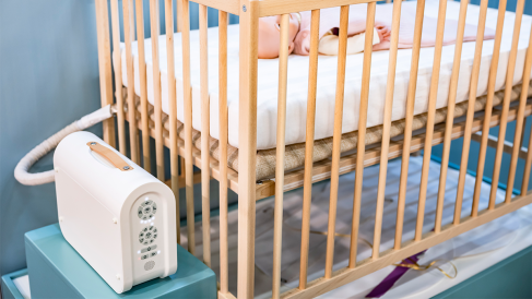 A wooden children's bed with a white mattress is shown, with a small person sleeping on it. Next to the bed, a pump is placed on a blue chest of drawers. The pump fills a mat to move the mattress. The room is bright and welcoming, suitable for a child's room.