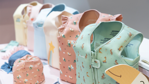 Various tops and small accessories are displayed on a white surface in pastel colors. The tops feature patterns such as stars, unicorns, glitter, and other playful designs. The presentation looks modern and inviting, as in an exhibition at a trade fair or shop.
