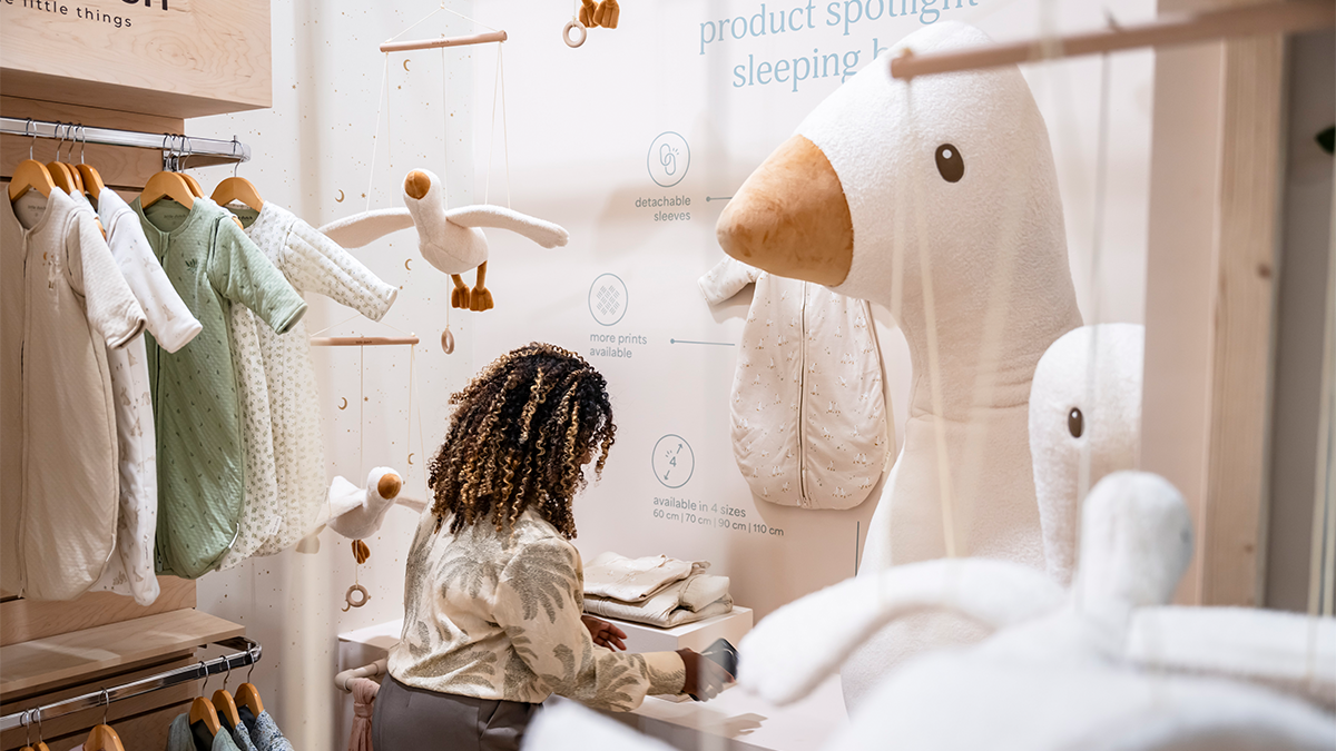 A retail display area features a wide selection of neutral and beige-colored baby and children's products presented in a store. The items—such as soft plush toys, small towels, blankets, pillows, fabric storage containers, baby toys, and decorative items like teddy bears and elephants—are attractively arranged on several white tables and shelves. In the background, several people can be seen exploring the shop or using their smartphones. The atmosphere is calm and inviting, ideal for shoppers looking for high-quality baby products.