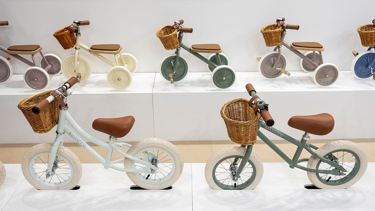 Four children's bicycles and a children's tricycle are displayed on white shelves against a white wall. The left bicycle is a light beige with black handles, seat, and tires. The center bicycle has a light blue pattern, gray handles, white tires, and a basket on the handlebar. The right bicycle is a light pink with patterns, wood-look handles, and white tires. At the bottom right is a small tricycle in light beige with black handles and tires. In front of the wall, two men are standing and looking at the bicycles while talking: one is wearing a gray polo shirt and has short gray hair, the other is wearing a white and blue striped shirt and has short dark hair. Their faces are blurred to protect privacy.