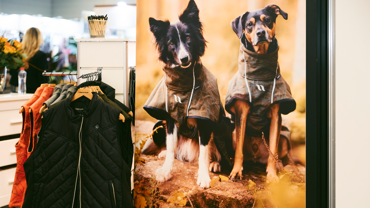 Two dogs wearing coats sit in front of a large poster showing the same dogs in coats
